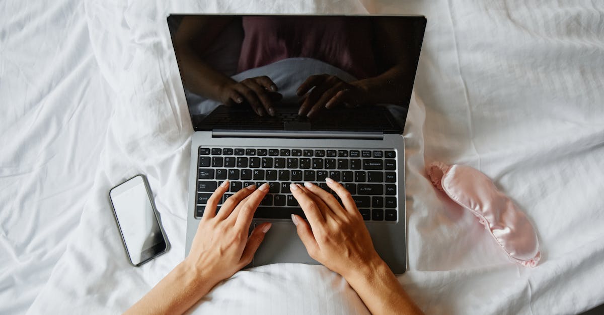 Person in bed researching on a laptop with a smartphone and sleep mask nearby